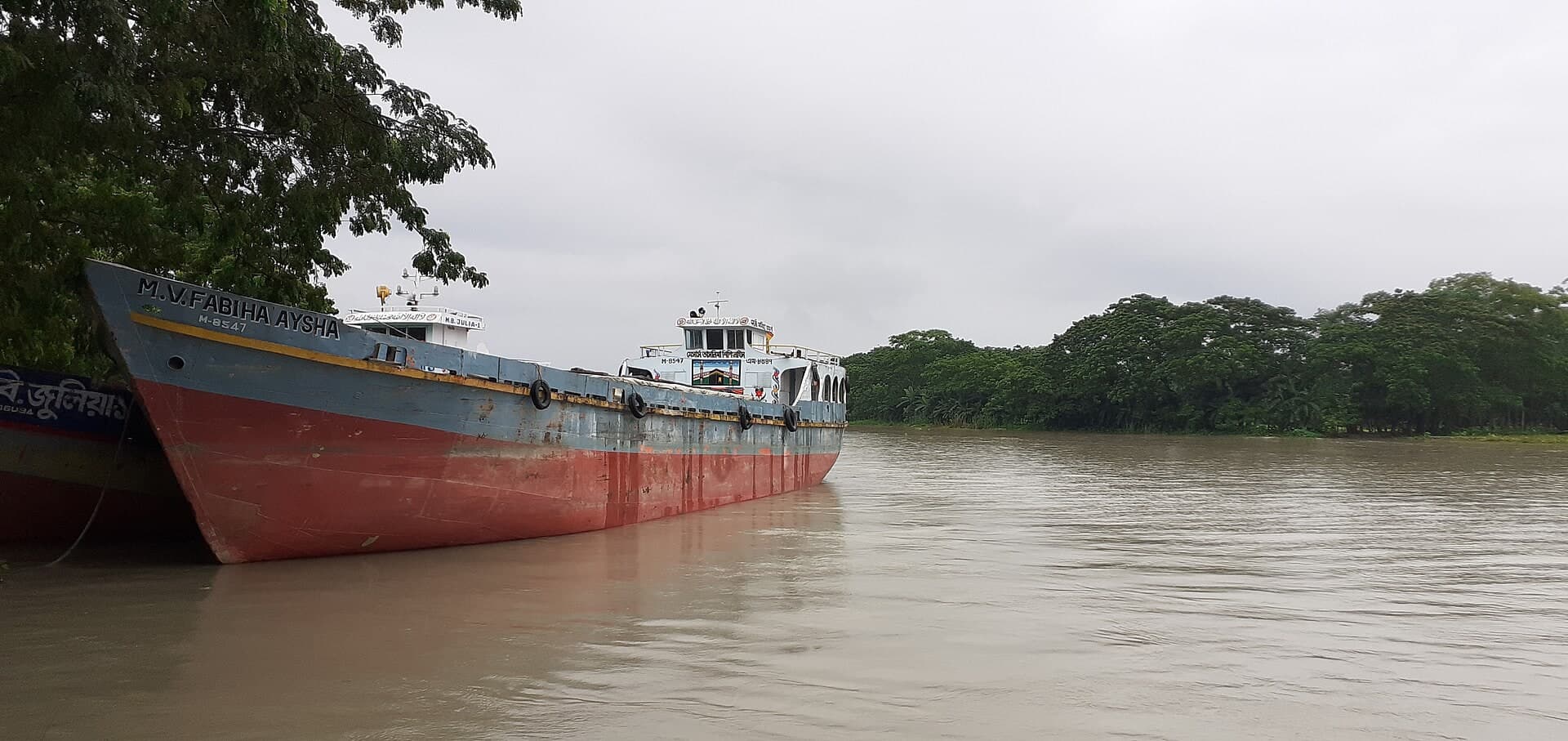 A red-hulled inland cargo vessel moored beside a tree-lined river bank.