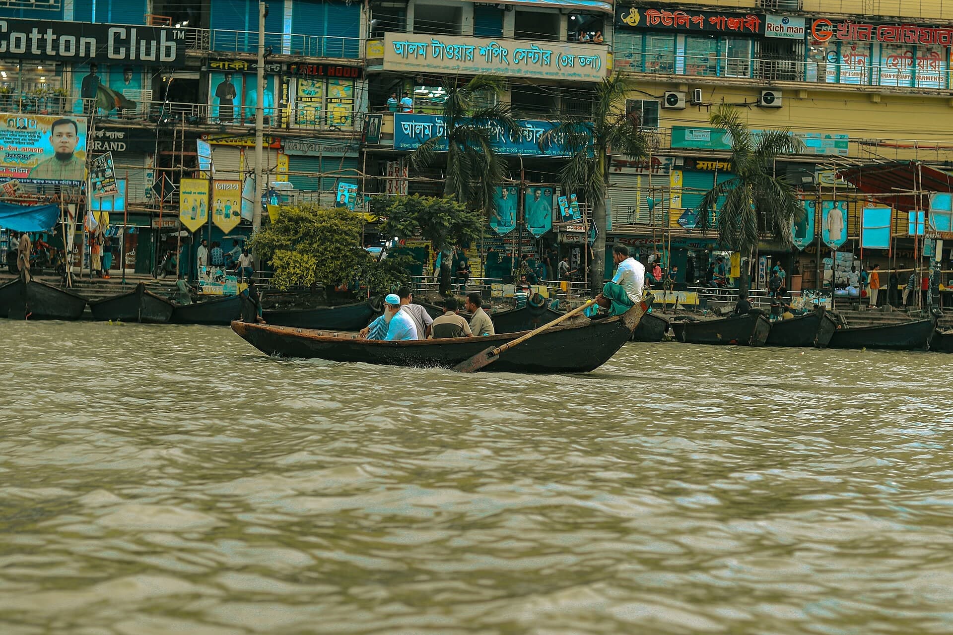 Wooden boats on the Buriganga in front of densely built riverside workshops and shops.
