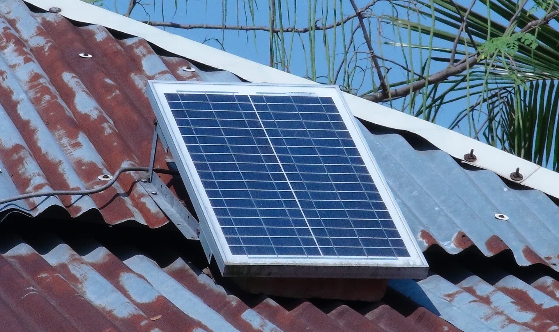 A small solar panel mounted on a corrugated metal house roof among palm leaves.