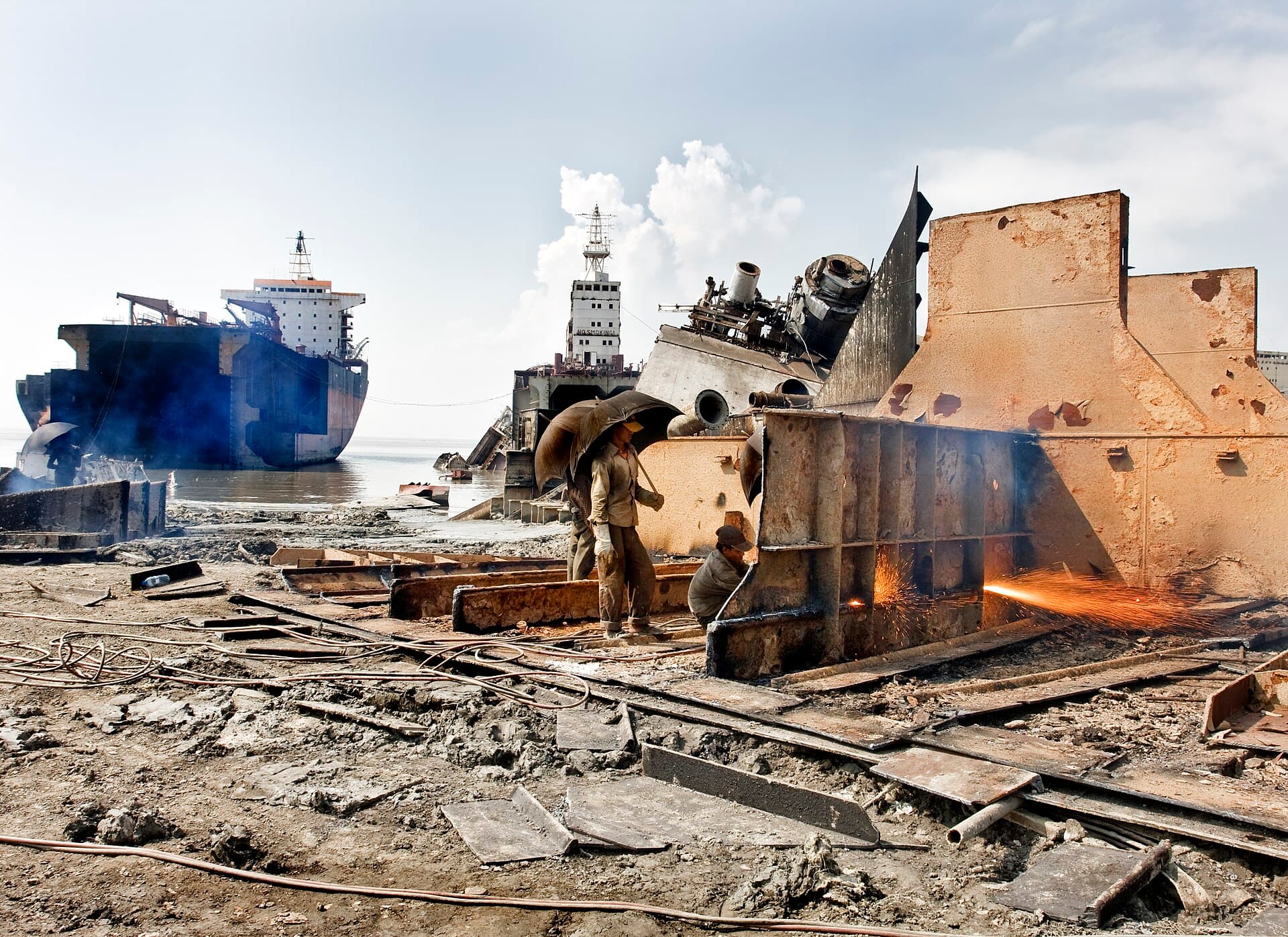Workers cutting a steel hull section with a gas torch at a ship-breaking yard, with beached ships behind them.