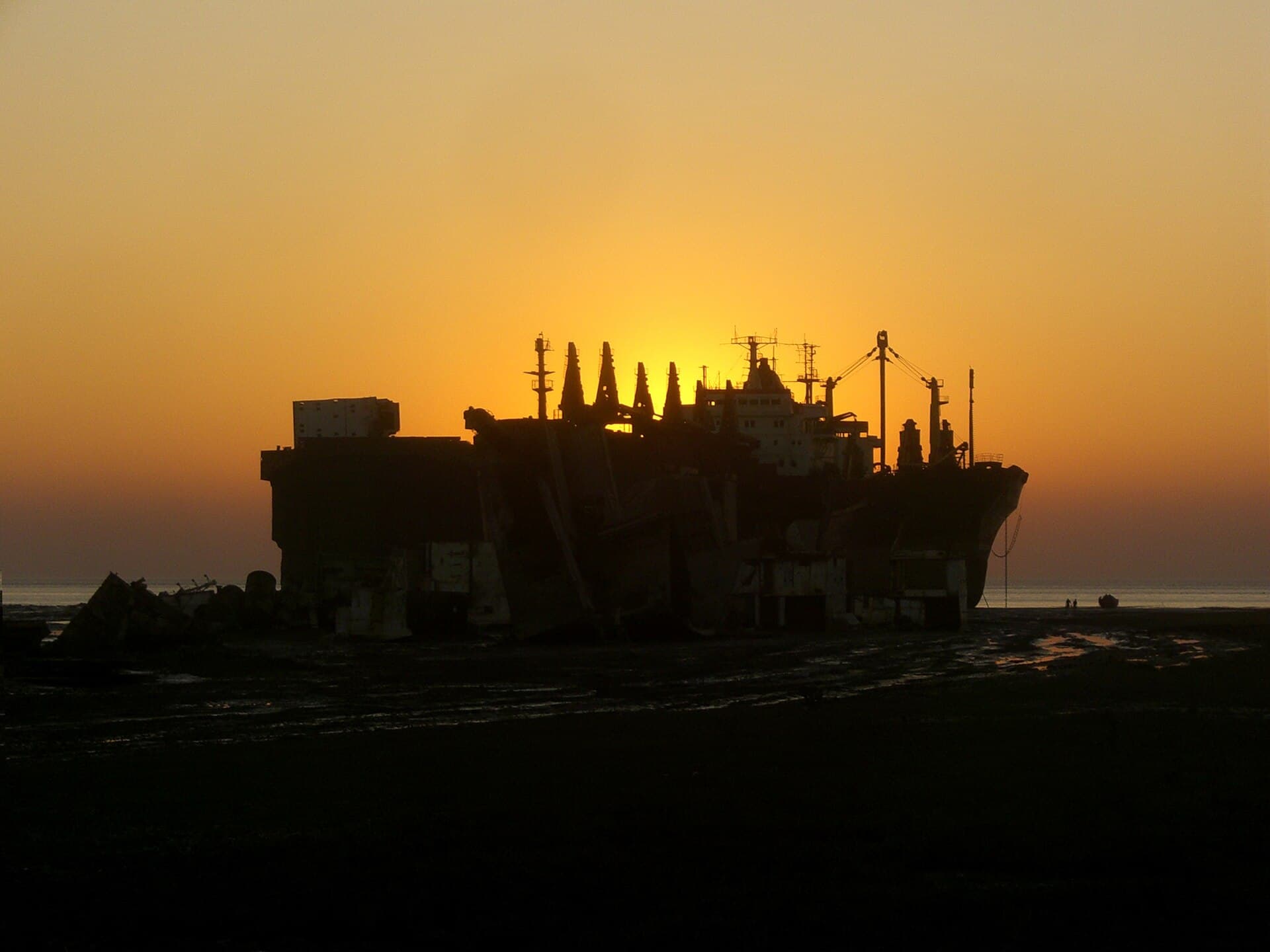 The silhouette of a partly dismantled ship on a tidal beach against an orange sunset.